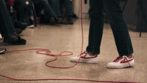 A person in jeans and white sneakers with red accents stands on a polished floor, holding a microphone with a long red cord. The cord is loosely coiled on the ground. Seated audience members are visible in the background.