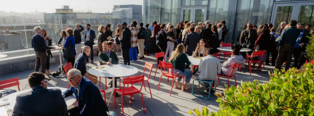 A large group of people mingles and converses on a sunny rooftop terrace with modern glass buildings in the background. Red chairs and tables are scattered around, and some greenery is visible in the foreground.
